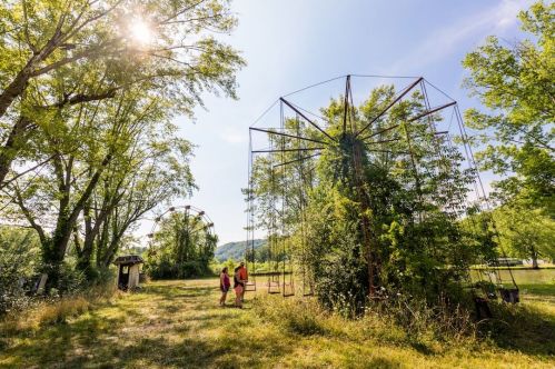Two people walk through a grassy area with overgrown amusement park rides and trees under a bright blue sky.