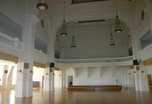 A spacious, empty hall with high ceilings, large windows, and wooden flooring, featuring elegant chandeliers.