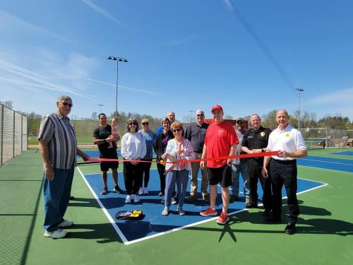 A group of people gathers on a tennis court for a ribbon-cutting ceremony, celebrating a new facility.