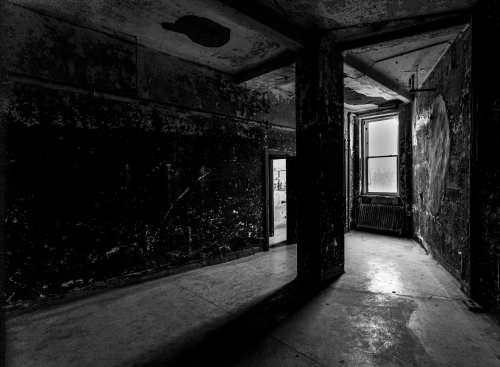 A dimly lit, abandoned room with peeling walls and a doorway leading to another space. Shadows create a haunting atmosphere.