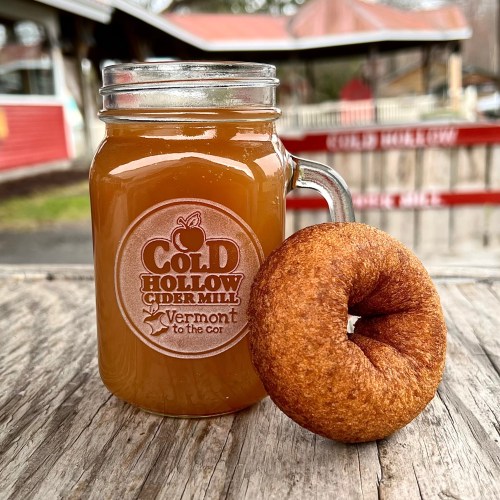 A mason jar of cider next to a cinnamon sugar donut on a wooden surface, with a rustic background.