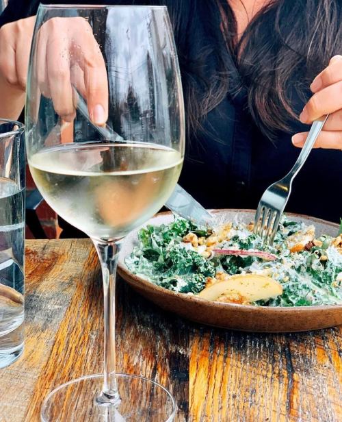 A close-up of a glass of white wine beside a person eating a salad with kale, apples, and nuts on a wooden table.