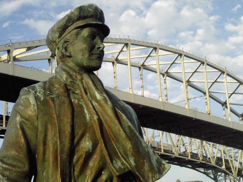 A bronze statue of a man in a cap and scarf, with a bridge and cloudy sky in the background.