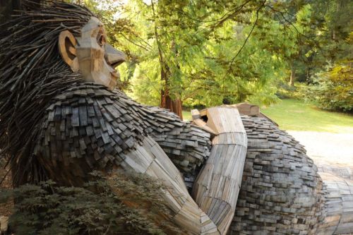 A large wooden sculpture of a figure with a textured body, surrounded by lush greenery in a serene outdoor setting.