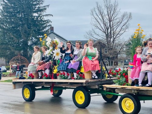 A group of women in colorful dresses wave from a decorated float during a parade, surrounded by flowers and greenery.