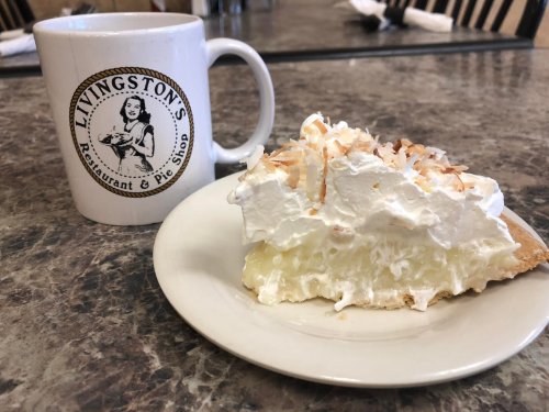 A slice of coconut cream pie with whipped cream on a plate, next to a coffee mug from Livingston's Restaurant & Pie Shop.