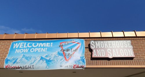 Banner reading "Welcome! Now Open!" with a mountain backdrop, next to a sign for "Smokehouse and Saloon."