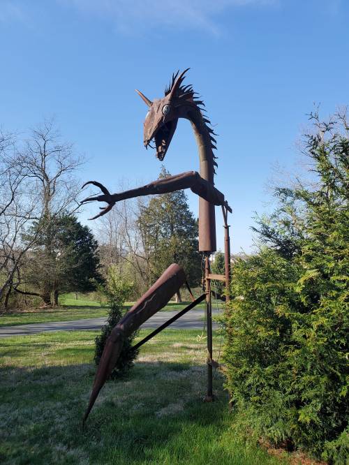 A large metal sculpture of a dragon-like creature stands among trees against a clear blue sky.