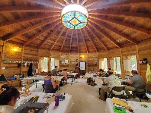 A group of people in a circular wooden room, engaged in a workshop with a presenter at the front and a stained glass ceiling.