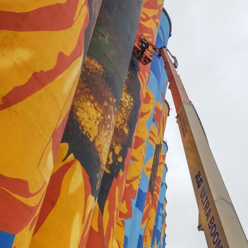 A worker on a lift paints a large mural on a colorful building facade against a cloudy sky.