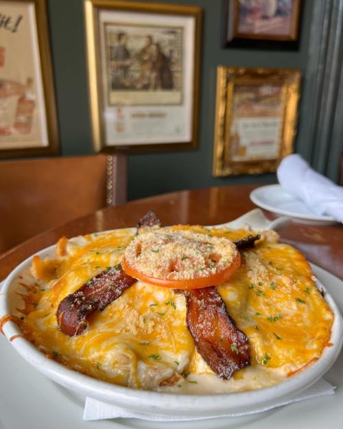 A creamy dish topped with melted cheese, bacon, and a slice of tomato, served in a bowl on a wooden table.