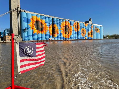 A vibrant mural of sunflowers on grain silos, with a flag in the foreground and water reflecting the blue sky.