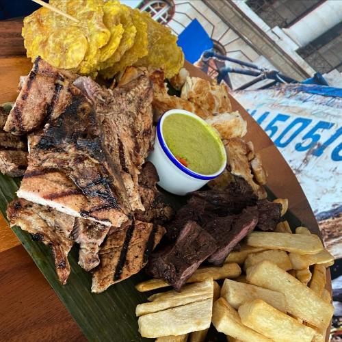 A vibrant platter of grilled meats, fried plantains, and cassava, served with a small bowl of green sauce.