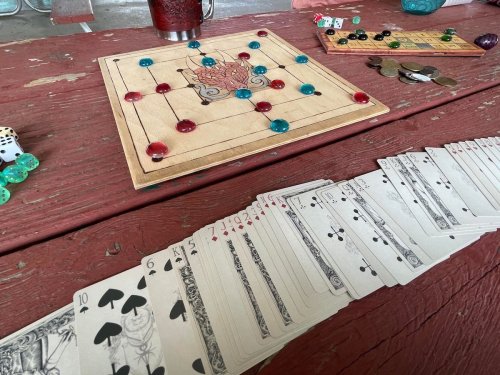 A wooden game board with colored pieces, dice, and a spread of playing cards on a red table.