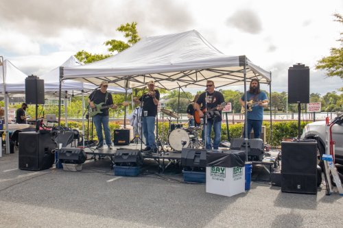 A band performs on an outdoor stage with instruments under a tent, surrounded by trees and parked cars.