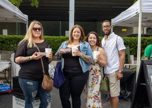 Four people stand together at an outdoor event, each holding a drink, smiling under a canopy.