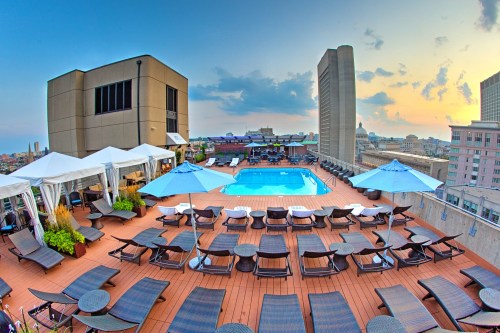 Rooftop pool area with lounge chairs and umbrellas, overlooking a city skyline at sunset.