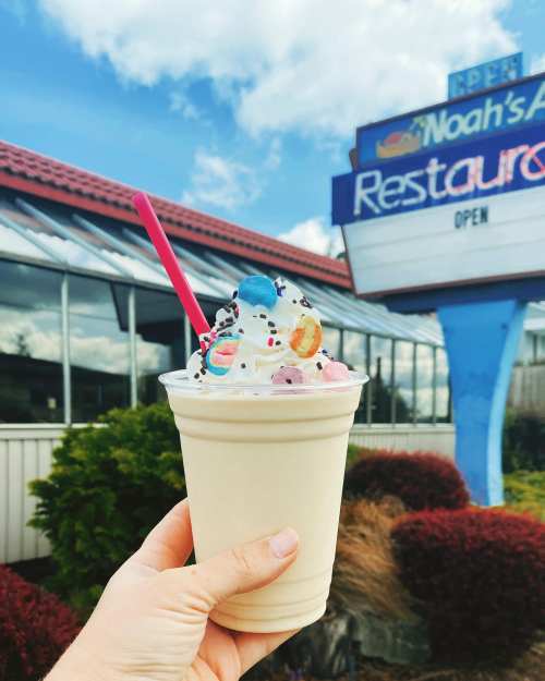 A hand holds a milkshake topped with whipped cream, sprinkles, and candy, in front of a restaurant sign.