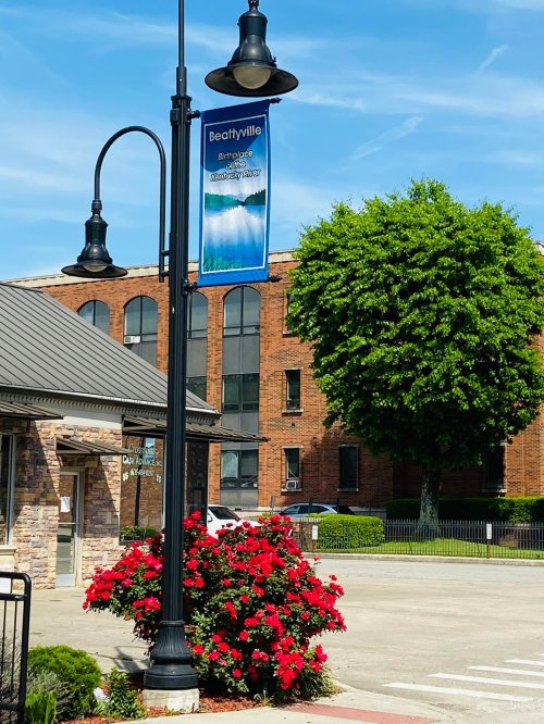 A street lamp with a banner reading "Beattyville" beside blooming red flowers and a green tree against a blue sky.