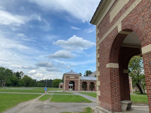 A person walks along a path near a brick archway, with green grass and a cloudy sky in the background.