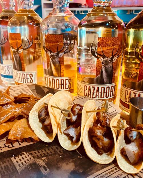 A platter of mini tacos filled with meat, accompanied by tortilla chips and bottles of Cazadores tequila in the background.