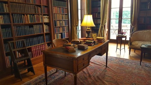 A cozy library with a wooden desk, stacked books, a lamp, and bookshelves filled with books in a sunlit room.