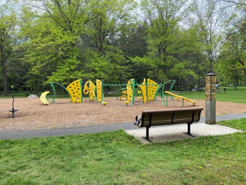 A playground with yellow climbing structures and green equipment, surrounded by trees and a bench nearby.