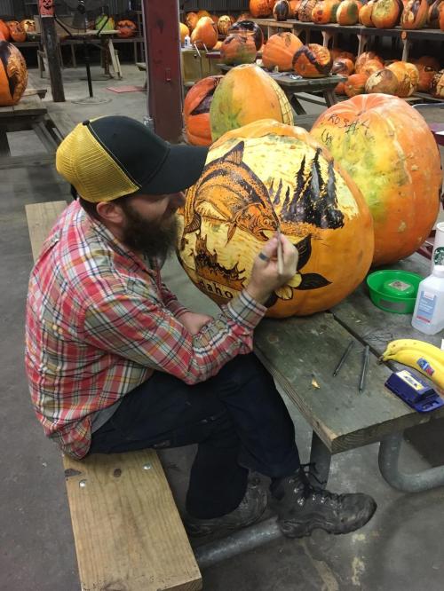 A person in a plaid shirt and hat carves a detailed design into a large pumpkin on a wooden table.