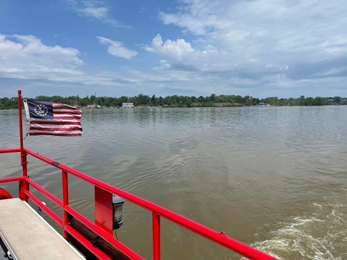 A view from a boat with a red railing, featuring an American flag and a calm river under a partly cloudy sky.