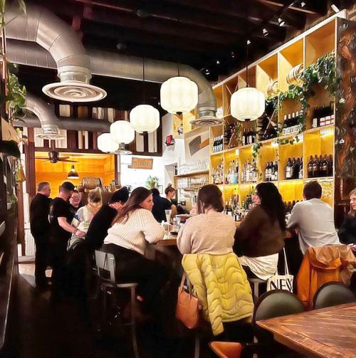 A lively restaurant interior with patrons at the bar, warm lighting, and shelves filled with bottles.