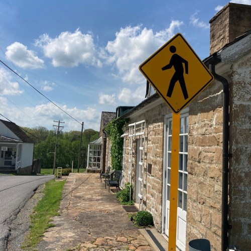 A pedestrian crossing sign stands beside a stone building along a quiet road under a blue sky with fluffy clouds.