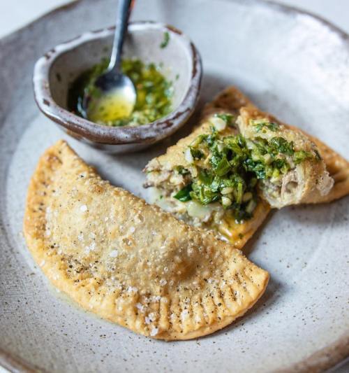 Two golden-brown empanadas on a plate, one cut open to reveal a green filling, alongside a small bowl of sauce.