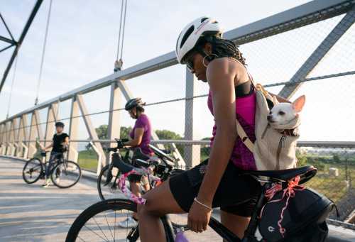 A cyclist with a small dog in a backpack pauses on a bridge, while two other cyclists ride nearby.