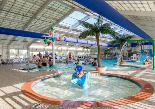 Indoor swimming pool with children playing, a slide, and palm trees, surrounded by lounge chairs and colorful decorations.