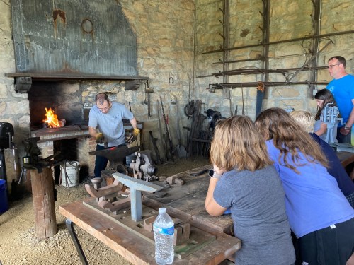 A blacksmith demonstrates his craft in a workshop, with onlookers observing and a fire burning in the forge.