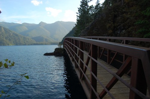A wooden bridge extends over calm water, surrounded by mountains and trees under a clear blue sky.