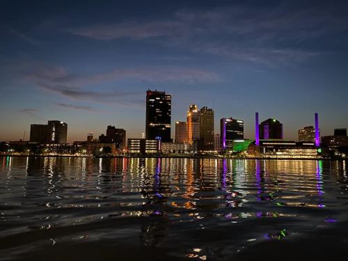 City skyline at dusk, reflecting on water, with illuminated buildings and a colorful glow in the sky.