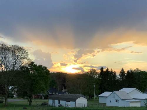 A sunset behind clouds, casting golden light over a rural landscape with trees and houses.