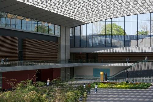 A modern indoor space with large windows, greenery, and people walking in a bright, open area.