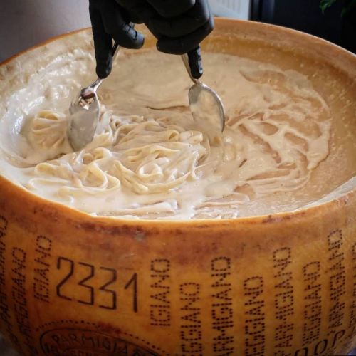 A hand using two spoons to mix pasta in a large wheel of cheese, creating a creamy dish.