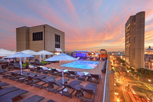 Rooftop pool with lounge chairs at sunset, overlooking a city skyline and busy streets below.