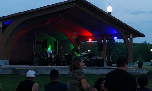 A band performs on stage at an outdoor venue, with colorful lights and an audience enjoying the music.