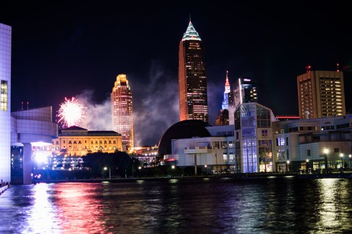 Cleveland skyline at night with fireworks, illuminated buildings, and reflections on the water.