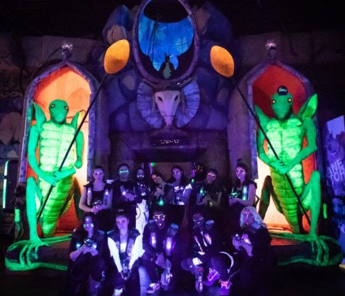 A group of people in futuristic outfits poses in front of glowing green figures and a colorful, surreal backdrop.