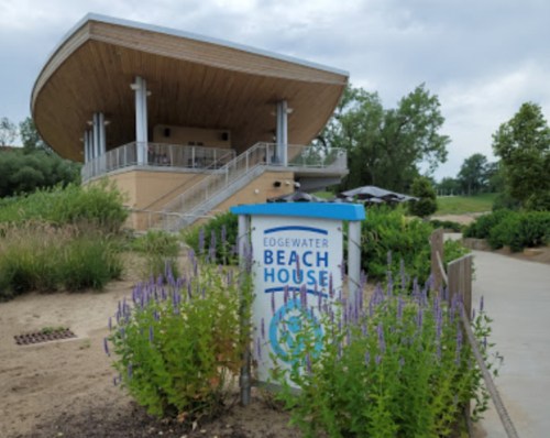 A modern beach house with a wooden roof, surrounded by greenery and purple flowers, near a sandy path.