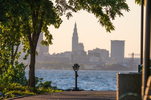 A scenic view of a city skyline across a lake, framed by trees and a viewing telescope in the foreground.
