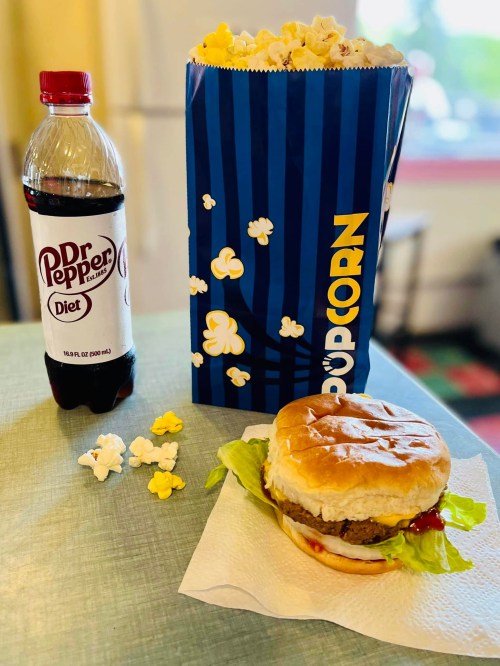A bottle of Diet Dr Pepper, a large popcorn box, and a burger with lettuce and ketchup on a napkin.