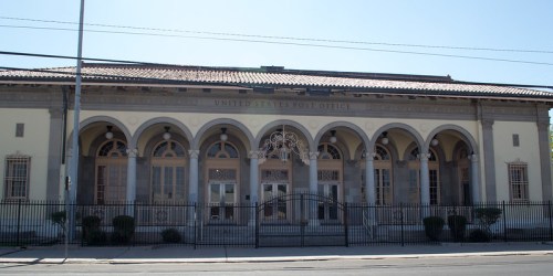 Historic United States Post Office building with arched windows and a gated entrance, set against a clear blue sky.