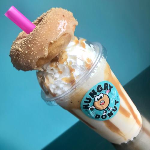 A creamy milkshake topped with whipped cream and a donut, featuring a pink straw and a "Hungry Donut" sticker.