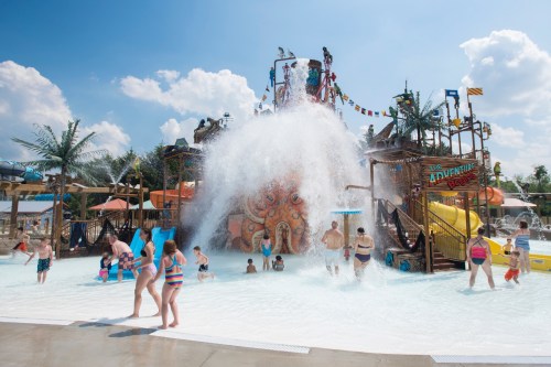 A vibrant water park scene with families enjoying water slides and a large splash area under a sunny sky.